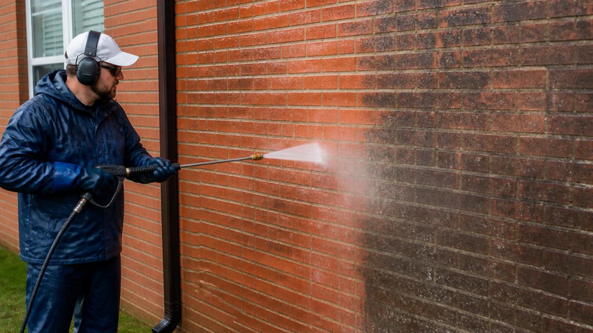A professional technician in safety gear using a low-pressure washer on a brick exterior wall, with water spraying gently and a clean section visible next to a dirty section